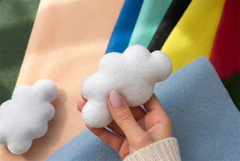 Hand holding a soft white felt cloud over colorful felt fabric sheets, showing the materials used for handmade baby mobiles and nursery decor.